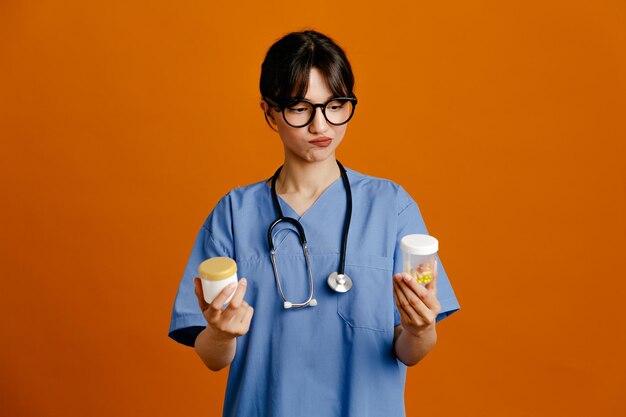 Contenitori portapillole scontenti giovane dottoressa che indossa uno stetoscopio fith uniforme isolato su sfondo arancione