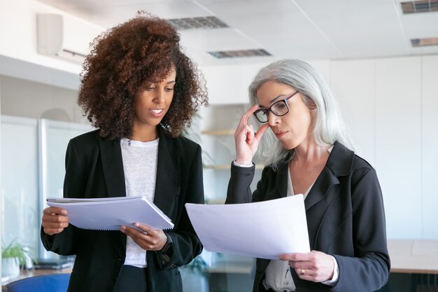 Content manager in bicchieri di lettura del documento con il giovane collega. Due imprenditrici di contenuto di successo che studiano i dati statistici e si incontrano nella stanza dell'ufficio. Concetto di lavoro di squadra, affari e gestione