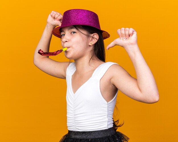 contenta giovane ragazza caucasica con cappello da festa viola che soffia fischio di festa e punta a se stessa isolata sulla parete arancione con spazio di copia