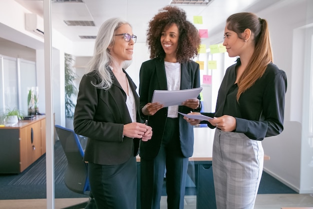 Contenti giovani donne di affari che discutono di statistiche e sorridono. Tre colleghe attraenti felici che stanno con i documenti e parlano nella sala conferenze. Concetto di lavoro di squadra, affari e gestione