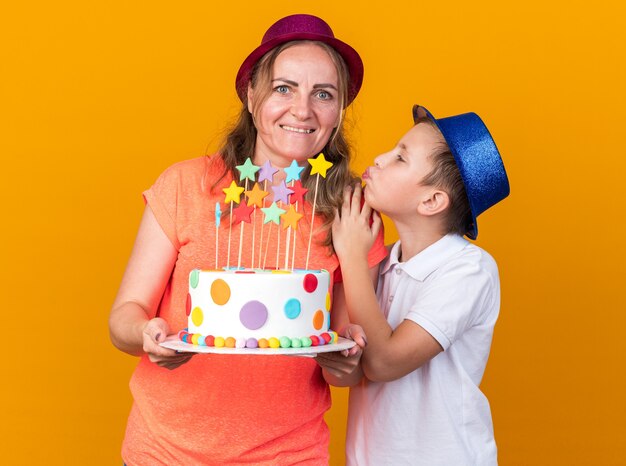 contento giovane ragazzo slavo con cappello da festa blu che cerca di baciare sua madre indossando cappello da festa viola e tenendo la torta di compleanno isolata sulla parete arancione con spazio di copia