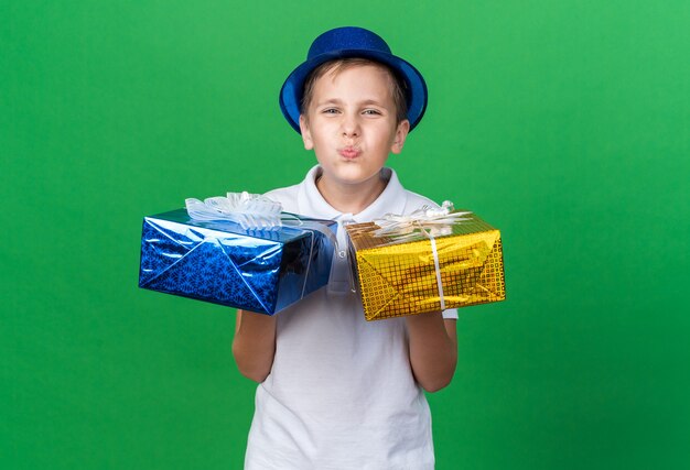 contento giovane ragazzo slavo con cappello da festa blu che tiene una scatola regalo su ogni mano isolata sulla parete verde con spazio copia