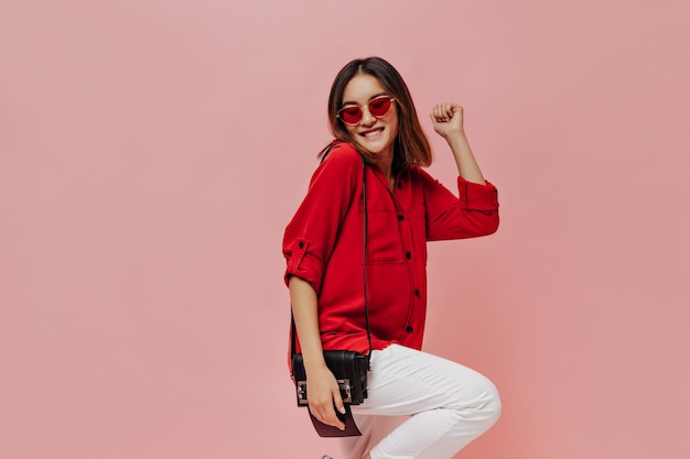 Cool giovane donna asiatica attraente in camicia rossa pantaloni bianchi e occhiali da sole sorrisi tiene borsetta e balli su sfondo rosa