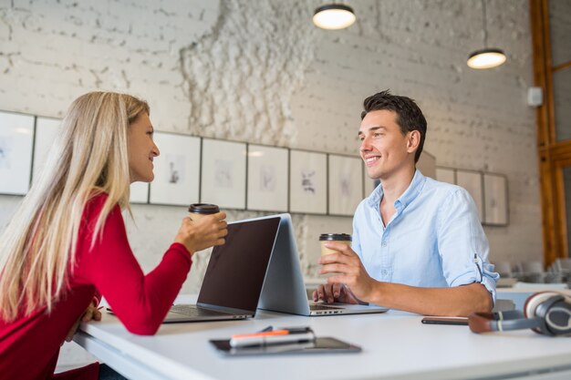 Cool giovane uomo e donna seduti al tavolo faccia a faccia, lavorando al computer portatile in ufficio di co-working