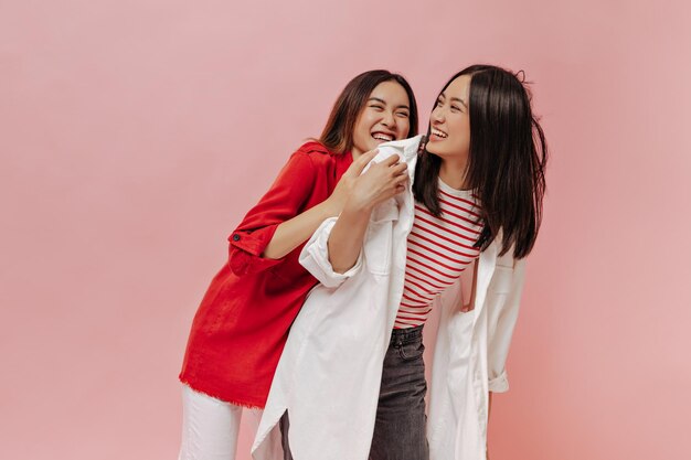 Cool giovani ragazze asiatiche di buon umore posano e si divertono su sfondo rosa Donna bruna in camicia lunga plaid e signora felice in camicetta rossa ridere su isolato