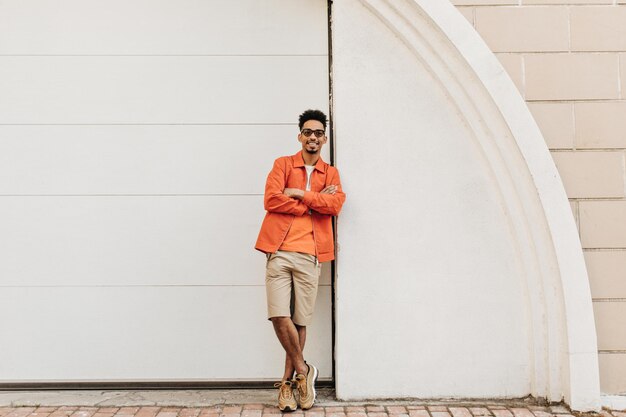 Cool uomo barbuto brunet in occhiali da sole pantaloncini beige e giacca arancione si appoggia sul muro bianco all'esterno e incrocia le braccia