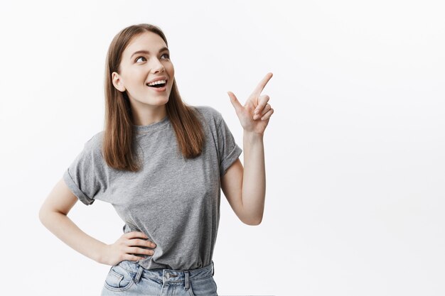 Copia spazio per pubblicità. Primo piano di una bella studentessa giovane indoeuropea con i capelli scuri in abiti grigi casual sorridendo brillantemente, guardando da parte con espressione felice, che punta sul retro bianco