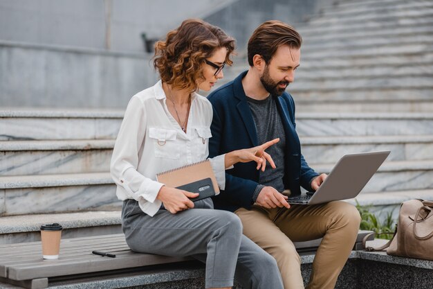 Coppia attraente di uomo e donna che parlano seduti sulle scale nel centro urbano, lavorando insieme sul laptop