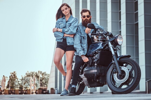 Coppia attraente hipster - maschio brutale barbuto in occhiali da sole e giacca di jeans seduto su una moto retrò e la sua giovane ragazza sensuale in piedi vicino, in posa contro il grattacielo.