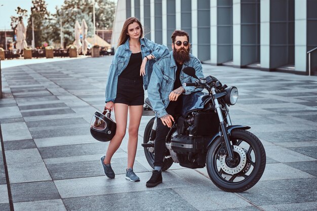 Coppia attraente hipster - maschio brutale barbuto in occhiali da sole e giacca di jeans seduto su una moto retrò e la sua giovane ragazza sensuale in piedi vicino, in posa contro il grattacielo.