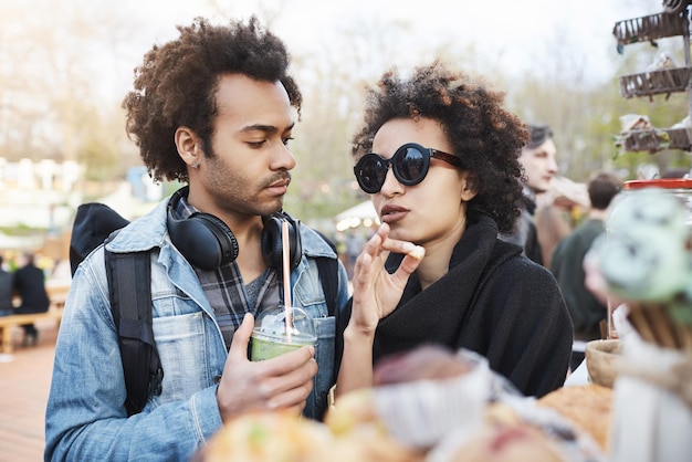 Coppia carina alla moda dalla carnagione scura in relazione con acconciature afro in piedi vicino al bancone del cibo nel parco raccogliendo qualcosa da mangiare e discutendo del gruppo che giocherà oggi