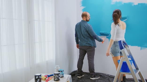 Coppia caucasica pittura parete con pennello a rullo e vernice blu. Ristrutturazione dell'appartamento e costruzione della casa durante la ristrutturazione e il miglioramento. Riparazione e decorazione.