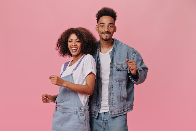 Coppia cool in abiti denim che ballano su sfondo rosa Donna riccia bruna in tuta e uomo in jeans sorridono isolato