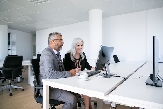 Coppia di allegri colleghi di lavoro guardando il contenuto sul monitor del computer, sorridendo e ridendo mentre era seduto al posto di lavoro. Concetto di comunicazione aziendale