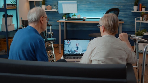 Coppia di anziani che usa la teleconferenza remota per parlare con il medico sul laptop, chiedendo informazioni sul trattamento. Persone anziane che discutono dell'assistenza sanitaria con il medico durante una teleconferenza online a casa.