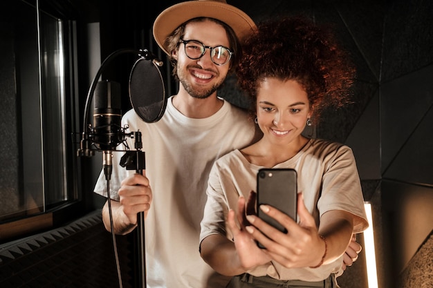 Coppia di attraenti cantanti alla moda che prendono felicemente selfie insieme sullo smartphone nel moderno studio di registrazione del suono