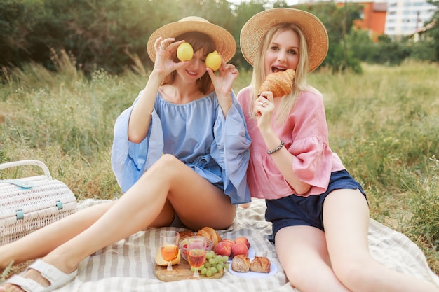 Coppia di belle donne graziose in posa sul prato nel parco estivo, gustando cibo gustoso, croissant e vino. Amici che godono del picnic.