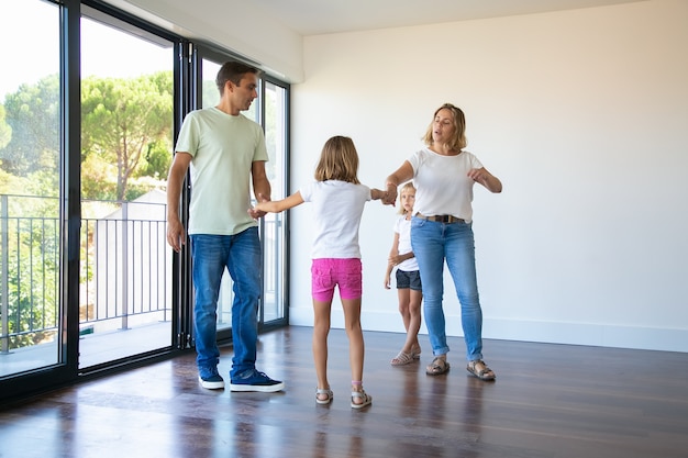 Coppia di genitori e due bambini che si godono la loro nuova casa, in piedi nella stanza vuota e tenendosi per mano, ballando