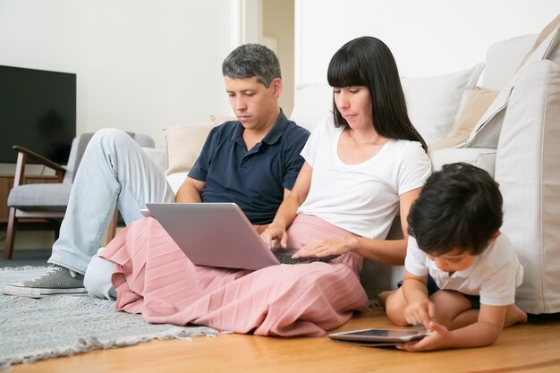 Coppia di genitori e figlio piccolo seduti insieme sul pavimento dell'appartamento, utilizzando tablet e laptop pc.