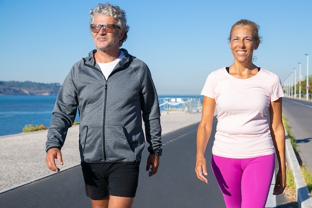 Coppia di jogging maturi camminando lungo la riva del fiume dopo la corsa mattutina. Vista frontale, colpo medio. Sport e concetto di pensionamento