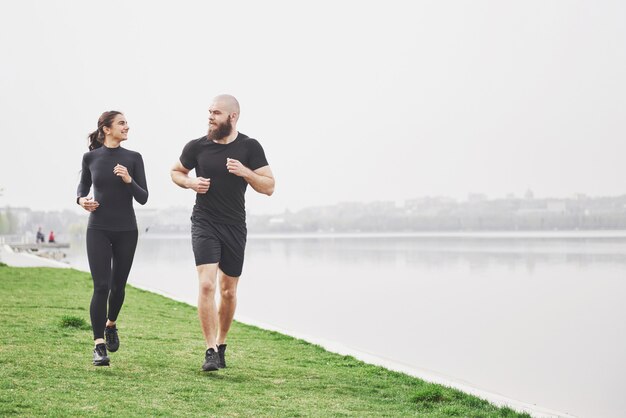 Coppia fare jogging e correre all'aperto nel parco vicino all'acqua. Giovane uomo e donna barbuti che si esercitano insieme nella mattina