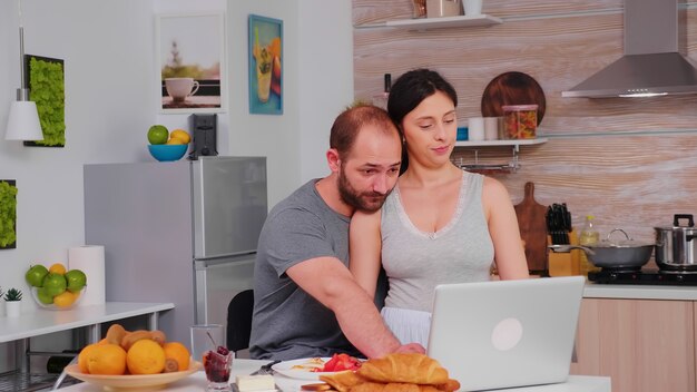 Coppia felice navigando su internet utilizzando il computer portatile durante la colazione in cucina. Marito e moglie sposati in pigiama che utilizzano la tecnologia moderna online di Internet, sorridenti e felici al mattino. leggendo bene