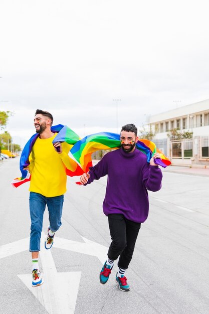 Coppia gay che corre lungo la strada tenendo le bandiere LGBT dietro le spalle