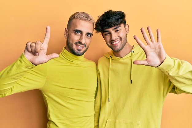 Coppia gay omosessuale in piedi insieme indossando abiti gialli che mostrano e puntano verso l'alto con le dita numero sette mentre sorridono fiduciosi e felici.