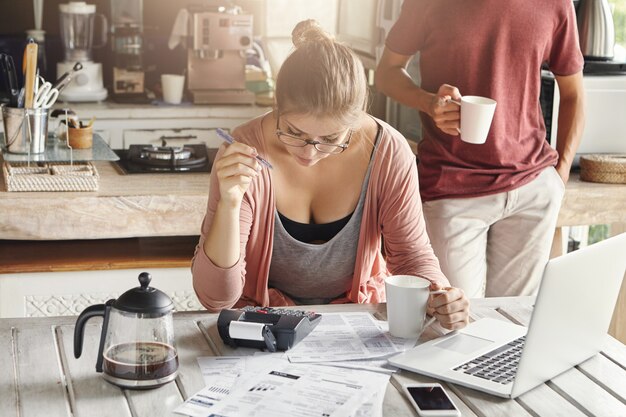 Coppia gestendo insieme il bilancio nazionale. Giovane femmina nella penna di tenuta di vetro mentre facendo i calcoli facendo uso del calcolatore e del computer portatile