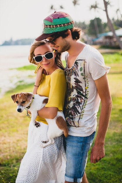 Coppia giovane hipster alla moda innamorata che tiene un cane al parco tropicale, sorridente e divertirsi durante le vacanze, indossando occhiali da sole, berretto, camicia gialla e stampata, romanticismo