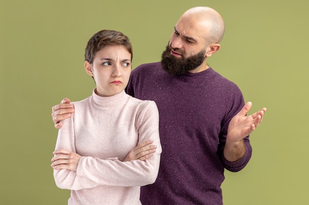 Coppia giovane in abiti casual uomo barbuto essendo confuso guardando la sua ragazza seria con i capelli corti il giorno di San Valentino concetto in piedi sopra il muro verde