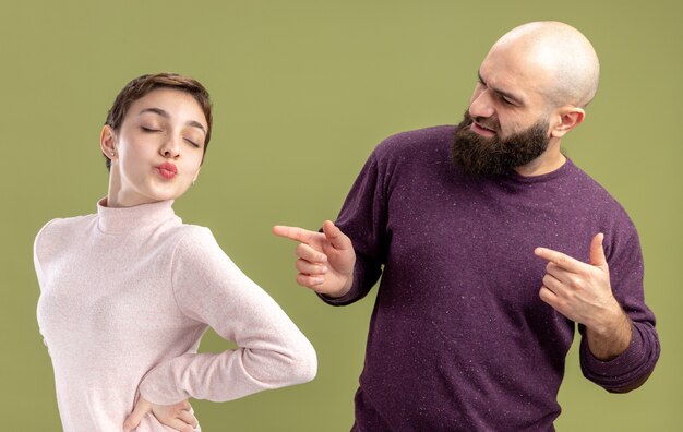 Coppia giovane in abiti casual uomo barbuto rivolto con il dito indice alla sua ragazza con i capelli corti per celebrare il giorno di San Valentino in piedi sopra il muro verde