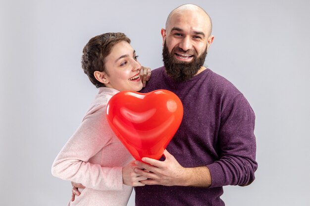 Coppia giovane in abiti casual uomo e donna azienda a forma di cuore palloncino sorridente allegramente felice in amore per celebrare il giorno di San Valentino in piedi su sfondo bianco