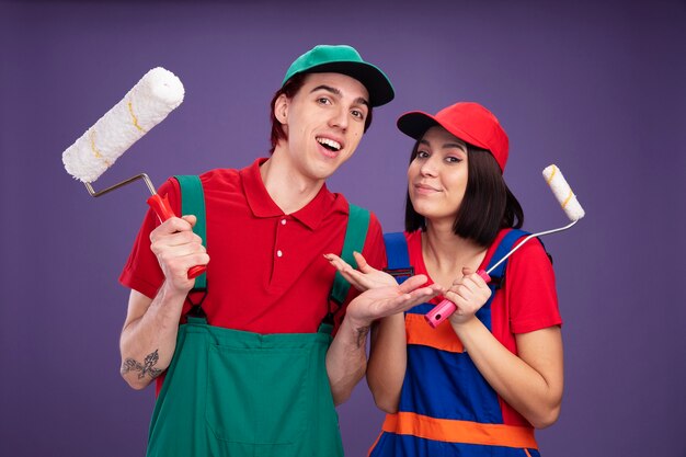 Coppia giovane ragazzo gioioso e ragazza compiaciuta in uniforme da operaio edile e cappuccio che tiene il rullo di vernice che mostra la mano vuota