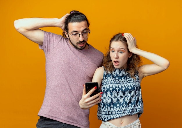 Coppia giovane uomo e donna con lo smartphone guardando sorpreso e confuso in piedi su sfondo arancione