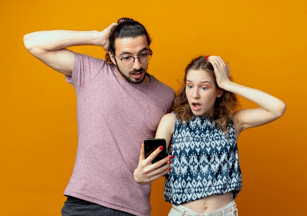 Coppia giovane uomo e donna con lo smartphone guardando sorpreso e confuso in piedi su sfondo arancione