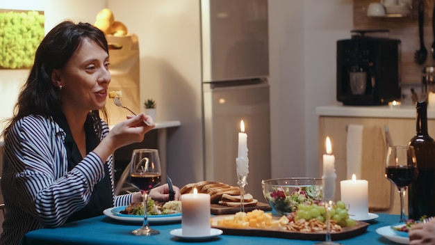 Coppia mangiare e bere vino con la donna in primo piano durante la cena festiva in cucina. Parlare felicemente seduti al tavolo della sala da pranzo, godersi il pasto a casa con momenti romantici a lume di candela