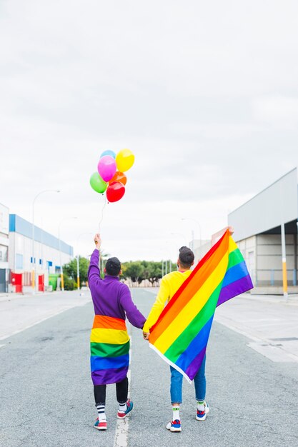 Coppia omosessuale che cammina lungo la strada tenendo palloncini e bandiere LGBT