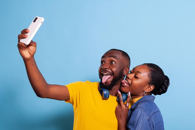 Coppia sciocca che tira fuori la lingua e scatta foto sullo smartphone, divertendosi con la fotografia. Uomo e donna sciocchi che scherzano con i selfie, creando ricordi sul cellulare.