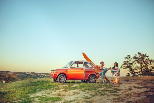 Coppia seduta e appoggiata sulla spiaggia a suonare la chitarra in una giornata estiva vicino al fiume. Amore, famiglia felice, vacanze, viaggi, concetto di estate.