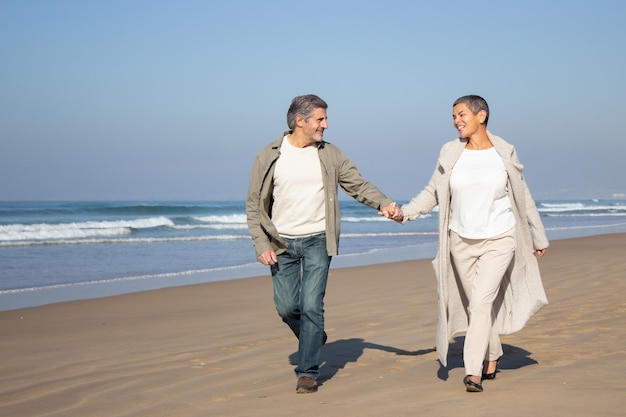 Coppia senior che cammina lungo la spiaggia in una giornata di sole. Uomo sorridente con i capelli grigi e felice donna dai capelli corti in cappotto che trascorrono del tempo insieme all'aperto. Relazione, concetto di partenariato