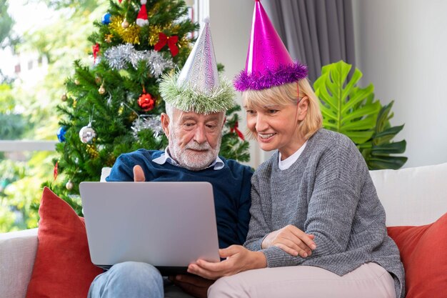 Coppia senior che usa il laptop per videochiamare per salutare la famiglia per il festival di Natale seduto sul divano con decorazioni e albero
