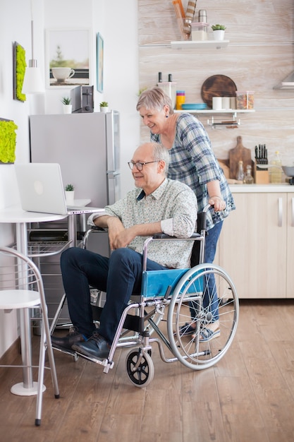 Coppia senior in una videochiamata in cucina. Uomo anziano disabile in sedia a rotelle e sua moglie che hanno una videoconferenza sul computer portatile in cucina. Un vecchio paralizzato e sua moglie hanno una conferenza online.