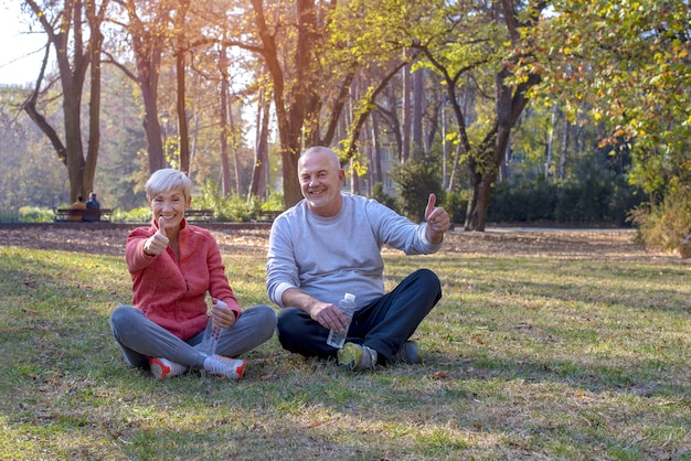 Coppia senior seduta sull'erba in un parco felicemente, alzando i pollici durante la luce del giorno