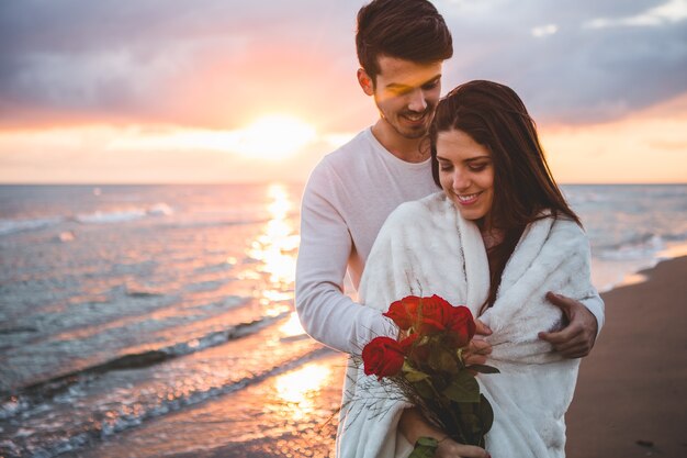 Coppia sorridente camminare sulla spiaggia con un mazzo di rose al tramonto