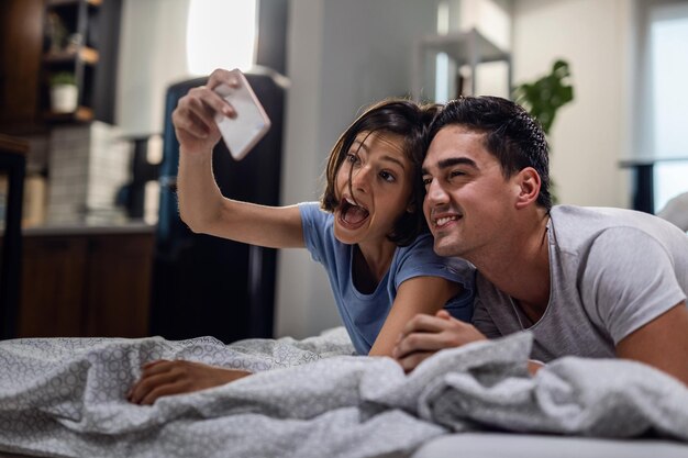 Coppia spensierata che si diverte insieme mentre fa facce e si fa selfie in camera da letto