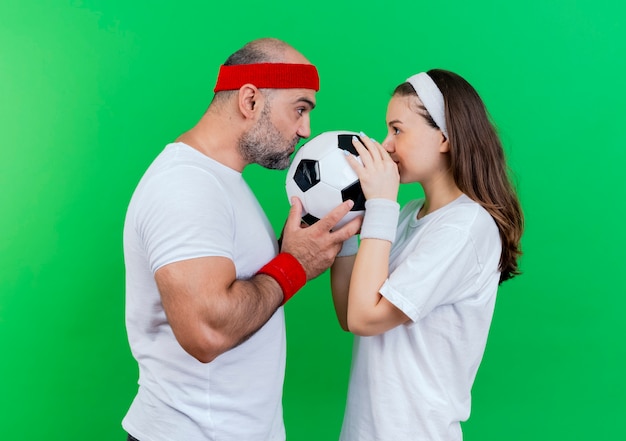 Coppia sportiva adulta che indossa fascia e braccialetti sia tenendo che baciando il pallone da calcio e guardandosi l'un l'altro