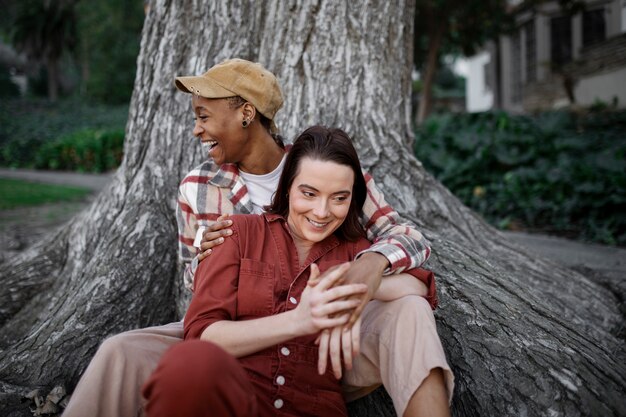 Coppia trans che si tiene per mano e sorride mentre si siede vicino a un grande albero al parco