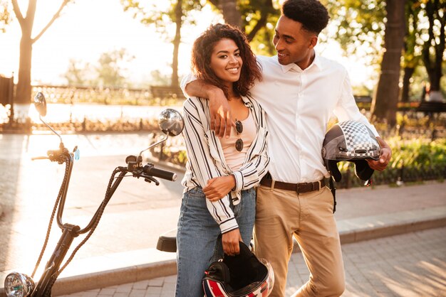 Coppie africane spensierate che abbracciano vicino alla motocicletta moderna nel parco