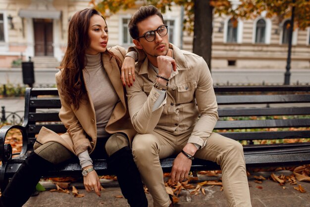 Coppie alla moda nella posa di amore all'aperto. Tendenze della moda autunnale. Modello del brunette con il ragazzo alla moda in cappotto beige che si siede sul banco.